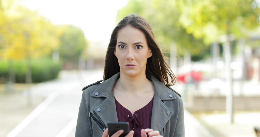 Front view of a perplexed woman walking checking smart phone content and then stops looking at camera in a park Royalty-Free Stock Footage #1019750350