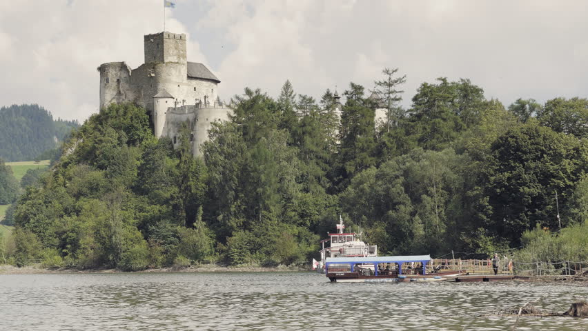 Medieval castle on hill above lake. Lake with ferry, tourist boat. Monumental stone historic building with large tower. Summer holiday scenery with clouds. Positive mood, beautiful landscape