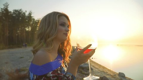 Cute young woman sitting on the shore of an amazing lake in the sunshine drinking wine from a glass 