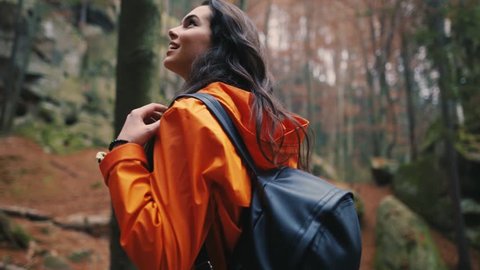 Charming brunette attractive young woman stand smile look around in forest outdoors with yellow leaves foliage autumn backpack hiker vacation portrait slow motion