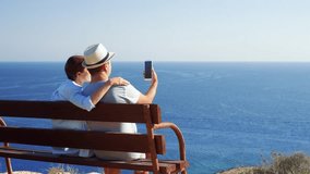 Young couple in love relaxing on bench at edge of cliff talking via messenger app having video chat on cellphone. Husband and wife on honeymoon enjoying view of Mediterranean sea using mobile phone