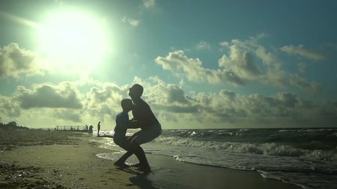 A young father walks with his son along the seashore. The sun shines brightly. Dad throws his son in the air. Family happiness. Trust relationship between father and son. Slow motion.