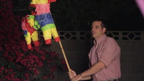 A man hits a brightly colored birthday piñata in a backyard at night.  The piñata flies out of the frame and gets caught on something. Camera flashes as he hits.