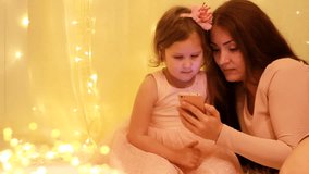 Mother and daughter watching video on smartphone on the background of lights boke.