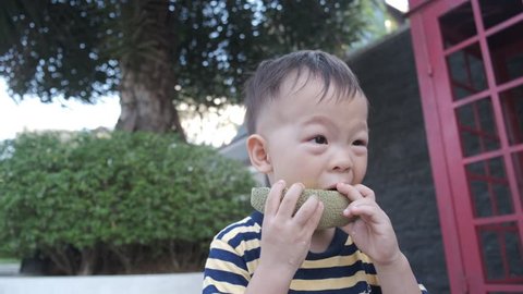 Cute Asian 2 years old toddler baby boy child standing at park in summer biting / eating fruit (melon) with his parent / father / dad (see only pointing hand), Healthy snacks & Self feeding concept