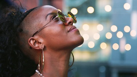 Young black woman looking up, turns to camera and takes off sunglasses, head shot, bokeh lights in background