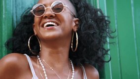 Young black woman wearing sunglasses leaning on green wall in the laughing, head and shoulders