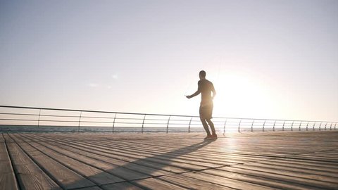 Motivated young athlete exercising with jumping rope at the seaside. Young man engaged in boxing working out outdoors. Slow motion. Sunrise. Steadicam
