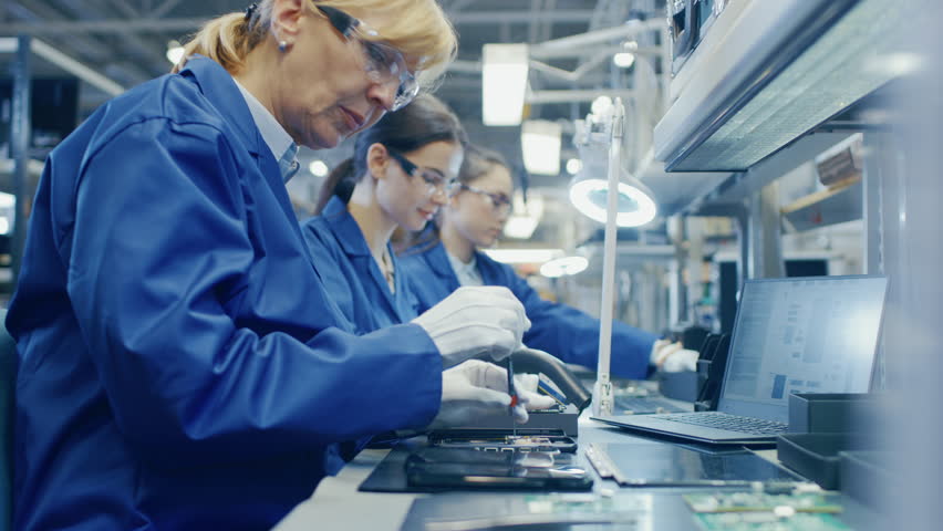 Woman Electronics Factory Worker in Blue Work Coat and Protective Glasses is Assembling Smartphones with Screwdriver. High Tech Factory Facility with more Employees in the Background. | Shutterstock HD Video #1019784358