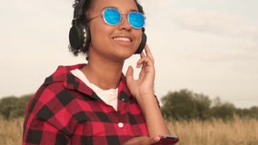 Slow motion tracking video clip of beautiful mixed race African American girl teenager young woman wearing blue sunglasses outdoors listening to music on wireless headphones 