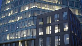 Modern multistory building facade, office center at dusk, windows with electric lighting and silhouettes of working people inside, side view, stock video