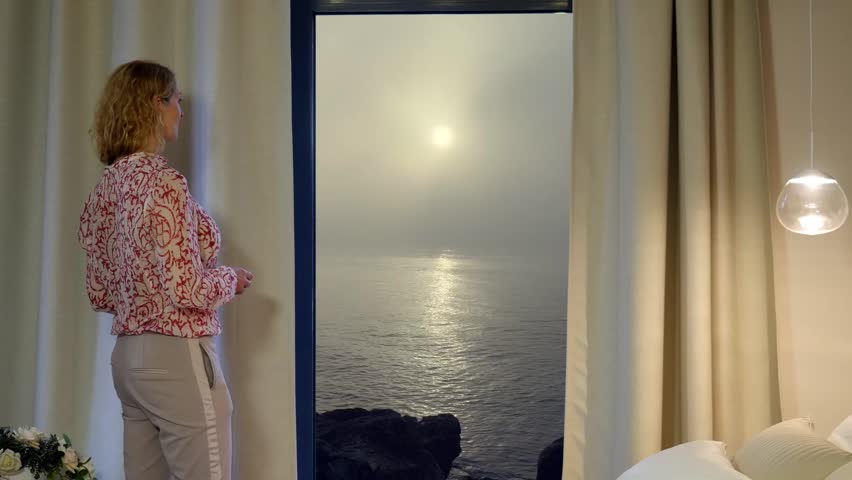 Beautiful woman stands at window drinks coffee and admires sunrise on Marbella bay in a misty morning, Back view, Full HD shot