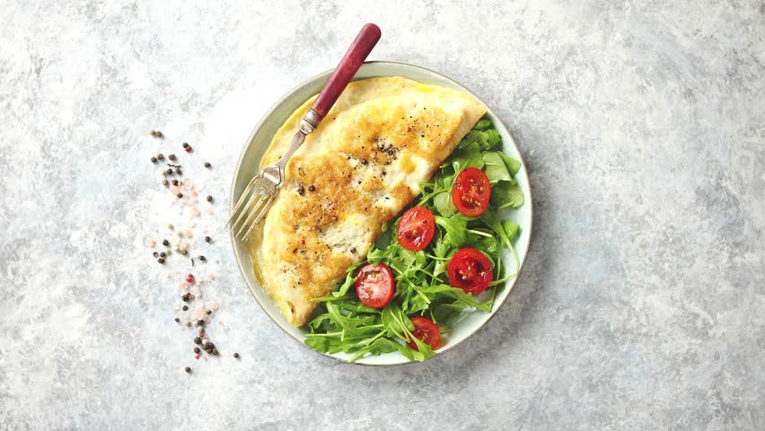 Classic egg omelette served with cherry tomato and arugula salad on side. Placed on white ceramic plate. Stone background with copy space.