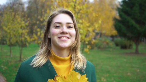 Attractive young woman walks along a bridge over a lake in an autumn park. Autumn portrait of beautiful woman over yellow leaves while walking in the park in fall. Blonde young girl in a green coat.