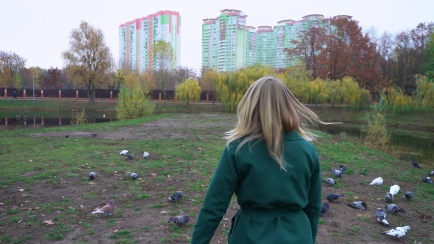Happy girl running in autumn park near the lake dispersing pigeons. Autumn portrait of beautiful woman over yellow leaves while walking in the park in fall. Blonde young girl in a green coat.