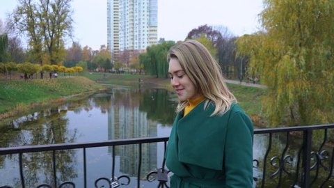 Attractive young woman walks along a bridge over a lake in an autumn park. Autumn portrait of beautiful woman over yellow leaves while walking in the park in fall. Blonde young girl in a green coat.