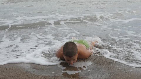 child lies on his stomach on the sandy shore in the sea waves. boy let the fountain of salt water out of his mouth. Kid washed sea waves.