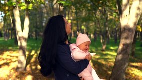 Beautiful young woman with long black hair standing outdoors in park and playing with little baby girl in pink suit, dancing and spinning for joy, smiling and looking at camera