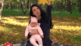Young woman with little baby sitting on the ground in park, throwing yellow leaves off her stretched hand