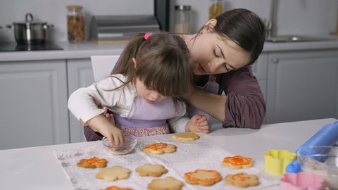 Concentrated little girl with down syndrome decorating gingerbread with icing in the kitchen. Caring mother teaching child with special needs baking and decorating homemade cookies with royal icing.