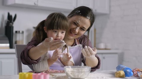 Carefree excited mother and joyful little girl with intellectual disability in matching apron, hands and faces stained with flour, having fun while enjoying cooking together in domestic kitchen.
