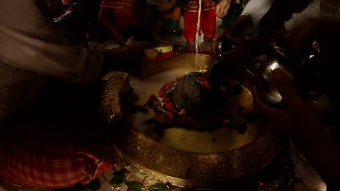 Rishikesh, Uttarakhand / India - July 30, 2018: hands do puja ritual prayer with candle to swayambhu natural lingam another people put on shivlinga flowers leaves of bilva and prasad pour water