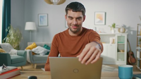 Confident Young Man Sitting at His Desk at Home, Opens and Starts Using Laptop. In the Background Living Room with Cozy Design.