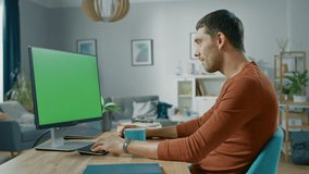 Handsome Smiling Man Sitting at His Desk at Home Uses Personal Computer with Mock-up Green Screen. He Lives in Cozy Aparment with Modern Living Room Interior.