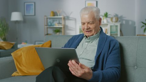Progressive Senior Man at Home Sitting on Sofa Uses Laptop Computer. Full of Life Elderly Man Relaxing at Home, Reading News on the Internet.