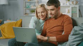 Cute Young Couple Sits on a Sofa at Home, They use Laptop Computer and Have Fun. Portrait of Beautiful Happy Young Couple in Love.