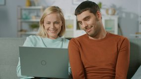 Cute Young Couple Sits on a Sofa at Home, They Look use Laptop Computer and Have Fun. Portrait of Beautiful Happy Young Couple in Love.