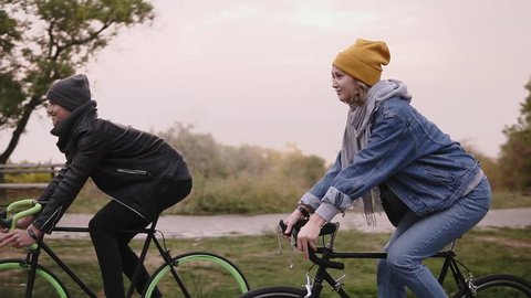 Young hipster couple enjoying cycling through park on trekking bikes. Two young people having great time together in autumn. Side view. Slow motion