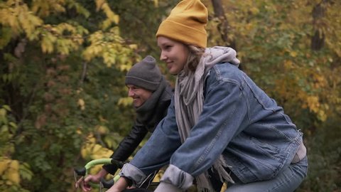 Happy, smiling friends or young couple cycling through the autumn park on bikes. Man and woman riding bikes together. Side view