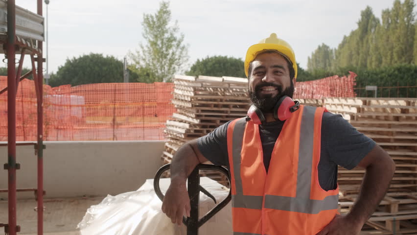 Skilled people working in construction site. Hispanic man at work in new house inside apartment building. Portrait of happy latino worker using manual forklift to move hardware and smiling at camera