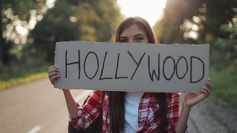 Young beautiful woman hitchhiking standing on the road holding Hollywood sign. Young sexy girl looking for a ride to start a journey. Summer time