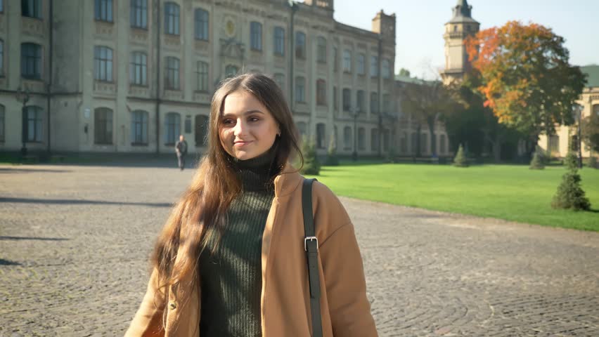 Happy caucasian woman is standing and enjoying sunshines on her face, nice weather, autumn vibes, casual stylish person standing, glancing at camera