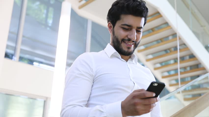 Multinational Bearded Man typing Massage on his Smarphone in Modern Office. Dreaming Startup Businessman Wearing Classical White Shirt. Looks Happy while Texting Funny Mails. Social Network. Startup.