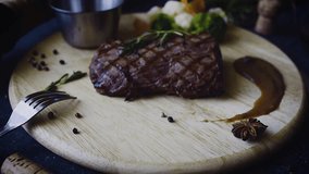 On the table there is a wooden textured Board with beef steak. Black Angus. Near vegetables, pepper, sauce. Red wine. Serving. Tableware. Restaurant. Video.