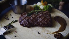 On the table there is a wooden textured Board with beef steak. Black Angus. Near vegetables, pepper, sauce. Red wine. Serving. Tableware. Restaurant. Black background. Video.