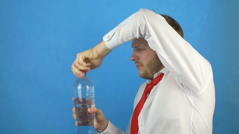A caucasian man with a beard in a white shirt and a hangover tie opens a bottle of mineral water and drinks, dipsomaniac, blue background