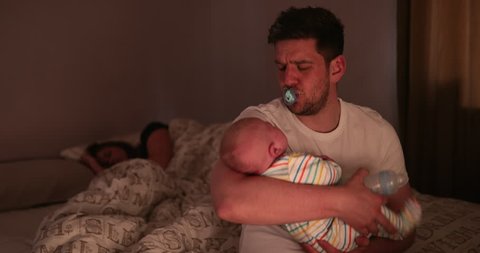 Dad checking on his newborn son, during the night, while his wife is fast asleep behind his on the same bed that he is sitting on to check his newborn.