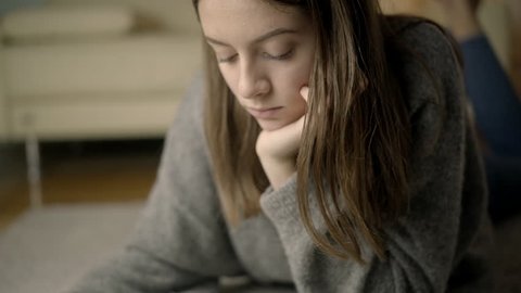 Young teen in a grey sweater, reading a book and studying for her exam in a cosy living room, while laying on the flor and sitting on a beige sofa, while being fully concentrated and looking in to the