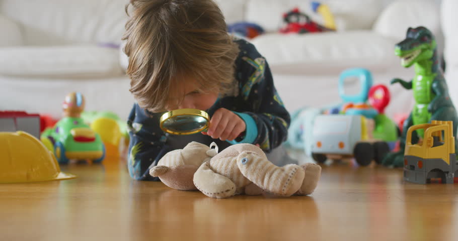 Cute little kid,  playing at home, in the living room, with toys around him, looking them with magnifying glass. 