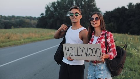 A young couple are hitchhiking standing on the road. A man and a woman stop the car on the highway with a sign Hollywood. Hitchhiking. Traveler. Transportation