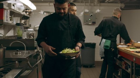 Group of professional chefs working at their stations at a long counter and a chef walking with a bowl of lettuce in industrial kitchen with soft lighting. Medium shot on 4k RED camera on a gimbal.