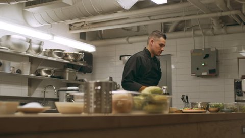 Professional chef garnishing wooden plate of salad and serves in industrial kitchen with soft lighting. Medium shot on 4k RED camera.