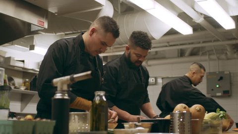 Group of professional chefs working at their stations at a long counter in industrial kitchen with soft lighting. Medium shot on 4k RED camera on a gimbal in slow motion.