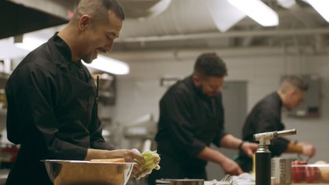 Professional chefs working at their stations at a long counter in industrial kitchen with soft lighting. Close up to medium shot on 4k RED camera on a gimbal in slow motion.
