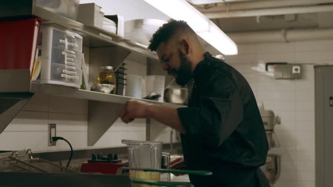 Professional chef bringing blender of orange puree to his work station to have other chefs taste in industrial kitchen with soft lighting. Medium shot on 4k RED camera on gimbal in slow motion,