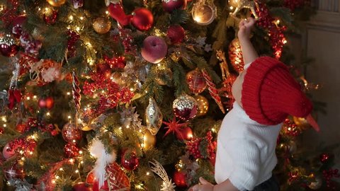 child in a white knitted sweater standing at the Christmas tree with garlands and considering toys
